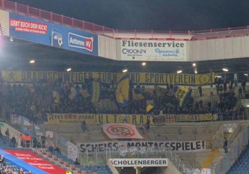 Eintracht Braunschweig-Fans hinter einem "SCHEISS MONTAGSSPIELE"-Banner im Gästeblock in Rostock. Dazu ein durchgestrichenes DFB-Wappen.