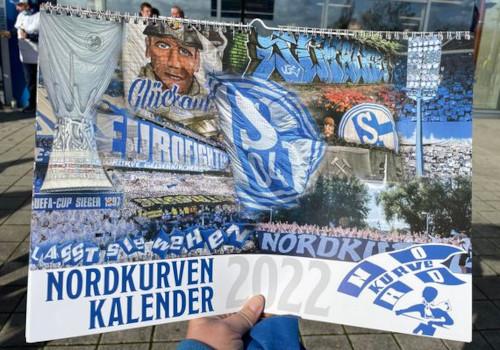 Der Nordkurvenkalender 2022 in der Hand gehalten vor der Arena auf Schalke.