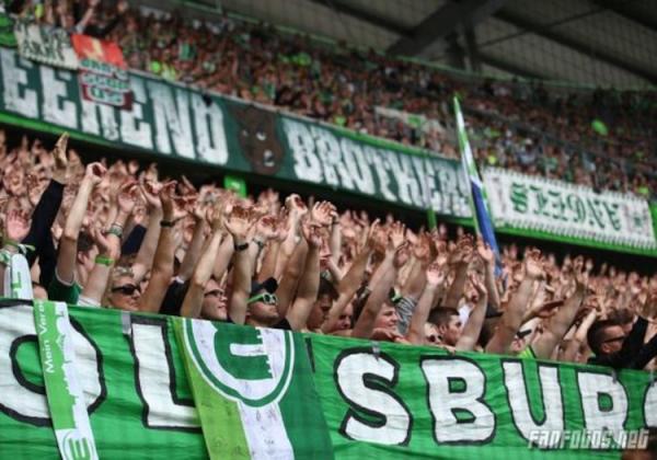 VfL Wolfsburg-Fans mit ausgestreckten Armen vor der Weekend Brothers-Zaunfahne.