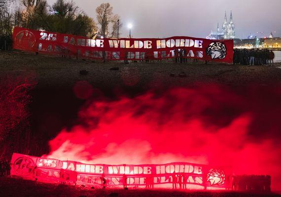 Gruppenfoto der Kölner Ultras aus anderen Gruppen hinter einem „25 Jahre Wilde Horde - Lang leben die Ultras“-Plakat auf der rechten Rheinseite.